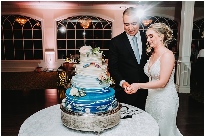 Professional Wedding Photo by Bokeh Love Photography, The Bradford Estate, bride and groom cutting the wedding cake that is designed with a beautiful white to blue ombre with Marlin, the fish on it, and lures